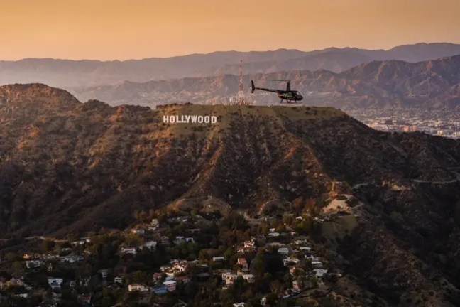 Helicopter flying over Hollywood sign with cityscape and mountains in background.